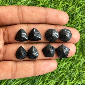 Puede incluir: Una colección de ocho piedras preciosas facetadas negras, que incluyen formas redondas, triangulares y pentagonales. Las piedras preciosas se muestran en una mano.