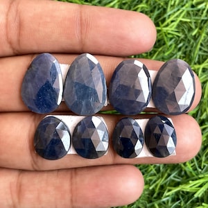 Puede incluir: Ocho zafiros facetados, de forma ovalada y color azul oscuro. Las piedras preciosas se presentan sobre una superficie blanca, sujetas por una mano, con hierba verde de fondo. Los zafiros tienen una superficie pulida y reflectante.