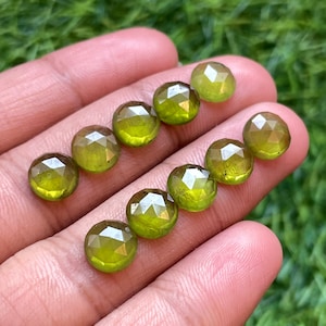 Puede incluir: Una colección de diez piedras preciosas verdes facetadas, probablemente peridotos, expuestas en una mano. Las piedras son redondas y tienen un acabado pulido.