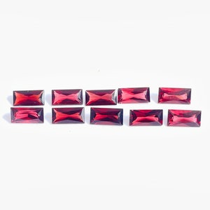 May include: Ten rectangular, faceted, deep red gemstones are arranged on a white surface. The gems have a baguette cut, with a rich, saturated color. The gemstones are evenly spaced, showcasing their clarity and brilliance. The overall presentation is clean and elegant.