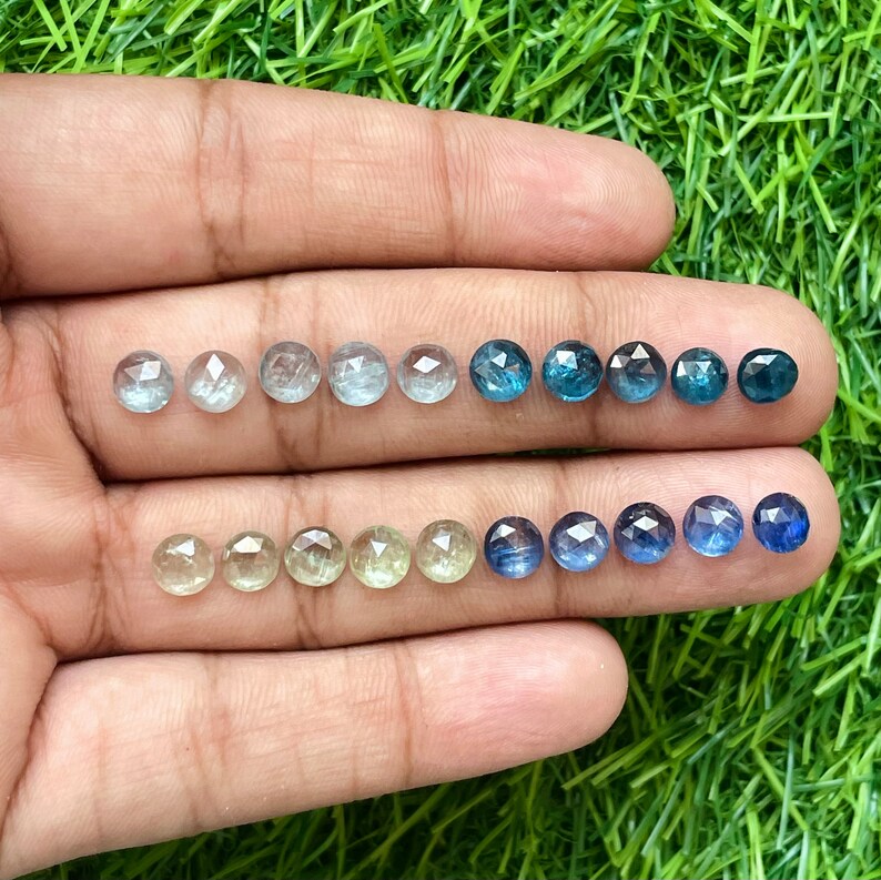 May include: A collection of round, faceted gemstones in various shades of blue and green. The stones are arranged in three rows on a hand, with the top row being the darkest blue and the bottom row being the lightest green.
