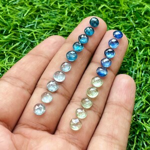 May include: A hand holding a variety of round, faceted gemstones in shades of blue and green. The gemstones are arranged in two rows, with the blue gemstones on the left and the green gemstones on the right. The gemstones are all approximately the same size and shape.