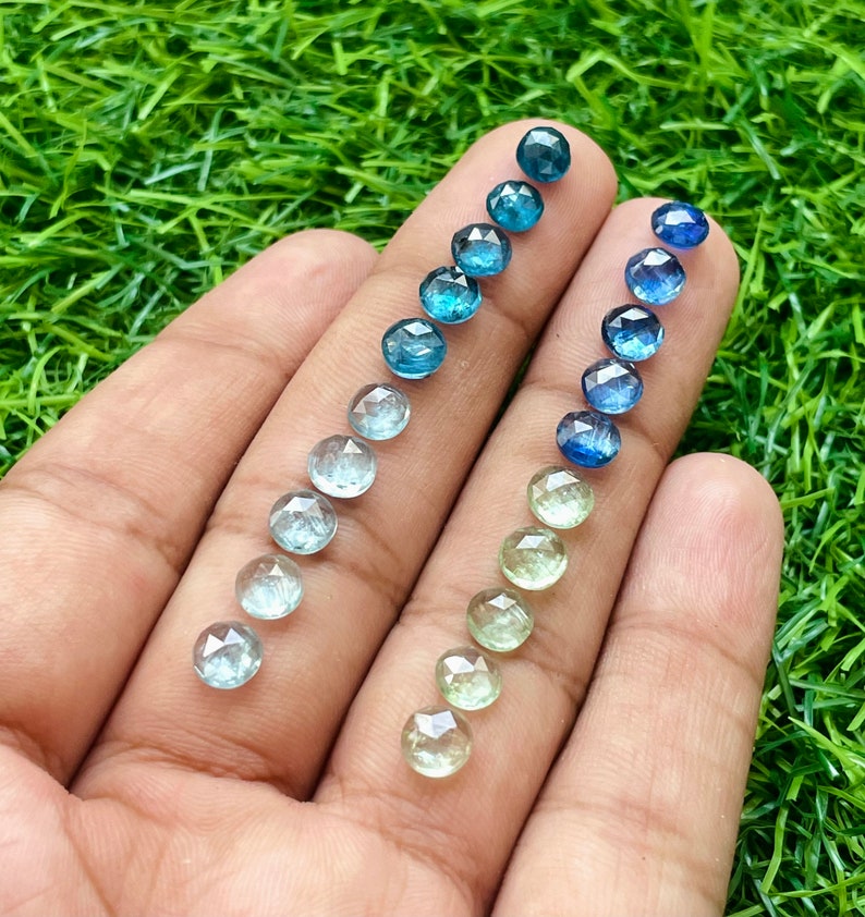 May include: A hand holding a variety of round, faceted gemstones in shades of blue and green. The gemstones are arranged in two rows, with the blue gemstones on the left and the green gemstones on the right. The gemstones are all approximately the same size and shape.