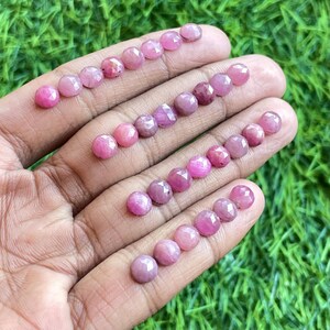 Pode incluir: Uma coleção de cabochões de rubi rosa, cada um com um corte facetado. As pedras estão dispostas em filas em uma mão, mostrando suas variações de tamanho e cor.
