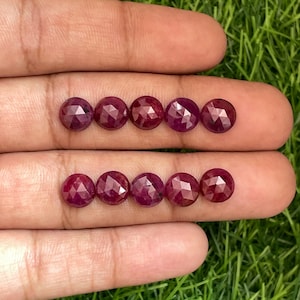 Puede incluir: Ocho piedras preciosas de rubí facetadas, cada una de un color rojo intenso, están dispuestas en dos filas de cuatro en una mano. Las piedras preciosas son redondas y tienen un acabado liso y pulido.