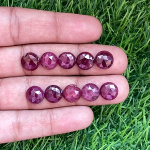 May include: Eight faceted ruby gemstones, each with a round shape and a deep red color. The gemstones are arranged in two rows of four, with the top row slightly offset from the bottom row.