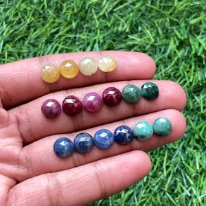 May include: Assortment of faceted gemstone cabochons in various colors, including yellow, red, blue, and green. The gemstones are arranged in rows on a hand.
