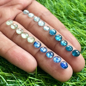 May include: A collection of round, faceted gemstones in various shades of blue and green. The stones are arranged in a gradient from light green to dark blue.