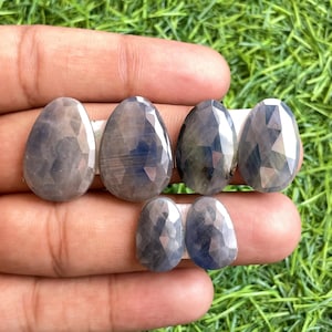 Puede incluir: Cinco piedras preciosas facetadas en forma de pera, de color azul oscuro. Las piedras preciosas están dispuestas en una fila en una mano.