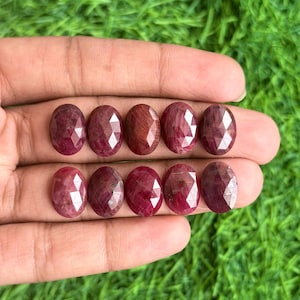 Puede incluir: Diez rubíes facetados de forma ovalada dispuestos en dos filas de cinco. Cada piedra preciosa muestra un tono rojo intenso con una superficie pulida de múltiples facetas que refleja la luz. Las piedras son uniformes en tamaño y forma.