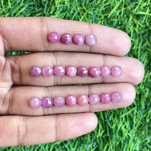 Puede incluir: Una colección de 20 piedras preciosas rosadas talladas en rosa, dispuestas en tres filas en una mano. Las piedras preciosas son facetadas y tienen un acabado brillante y pulido.