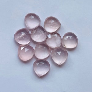Könnte beinhalten: Eine Gruppe von zehn facettierten Rosenquarzsteinen in Tropfenform. Die Steine sind hellrosa und haben eine durchscheinende Optik.