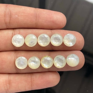 Puede incluir: Se muestran diez piedras preciosas redondas, facetadas y blanquecinas. Las gemas tienen un brillo nacarado y están dispuestas en dos filas de cinco. Las facetas captan la luz, creando un efecto brillante. Estas piedras podrían usarse para joyería.