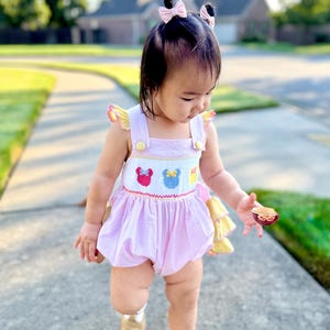 May include: A baby wearing a pink and white gingham romper with embroidered cartoon character faces. The romper has yellow accents and is paired with gold shoes. The baby has pink bows in their hair.