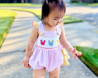 May include: A baby wearing a pink and white gingham romper with embroidered cartoon character faces. The romper has yellow accents and is paired with gold shoes. The baby has pink bows in their hair.