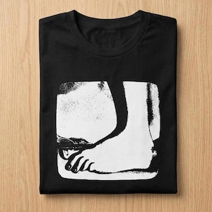 May include: Black t-shirt with a black and white graphic of a foot and ankle. The graphic is within a white square. The t-shirt is folded on a light brown wooden surface.