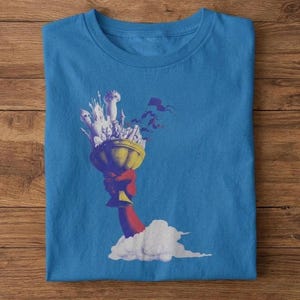 Puede incluir: Camiseta azul con una ilustración surrealista. Un cáliz dorado es sostenido por un brazo rojo, con un castillo blanco y banderas volando encima. Una nube blanca está en la base del brazo.