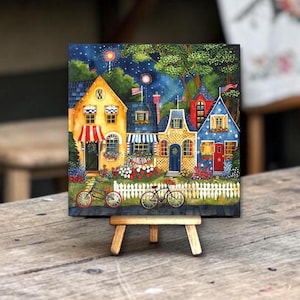 May include: A colorful painting of a row of houses with bicycles in front, displayed on a small wooden easel. The houses are painted in various colors, with details like awnings, flags, and flower boxes. The background shows a night sky with fireworks.
