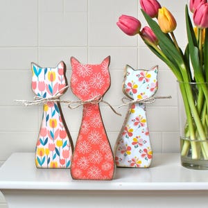 May include: Three wooden cat figurines with floral patterns in pink, white, and yellow. Each cat is tied with twine and stands on a white surface. A vase of pink and yellow tulips is to the right.