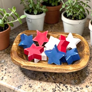 May include: A wooden bowl filled with red, white, and blue star-shaped decorations. The bowl sits on a speckled countertop, with small potted plants in the background. The stars are likely for patriotic or holiday decor.