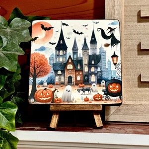 May include: A Halloween-themed square art print on a small wooden easel. The print depicts a spooky haunted house, pumpkins, ghosts, bats, and a witch flying on a broomstick. The color palette includes orange, black, and blue.