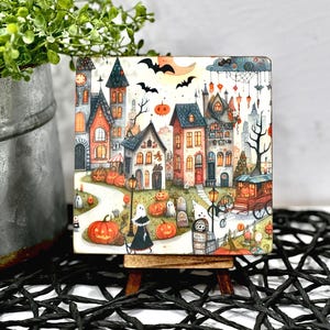 May include: A square Halloween-themed artwork featuring a whimsical town scene with colorful houses, pumpkins, ghosts, and bats. The artwork is displayed on a small wooden easel, perfect for seasonal decor.
