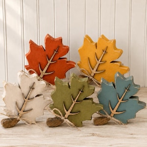 May include: Five wooden leaf-shaped decorations in orange, yellow, white, olive green, and blue. Each leaf features a cutout wooden vein detail attached to the center and is tied with a small piece of twine at the base of the stem.