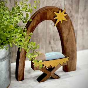 Op de afbeelding: Een houten kerststal met een donkerbruine boog en een gouden ster. Binnen de boog staat een wieg met een lichtblauw kussen en een houten baby. De wieg wordt ondersteund door een houten standaard. Een groene plant in een metalen pot staat op de achtergrond.