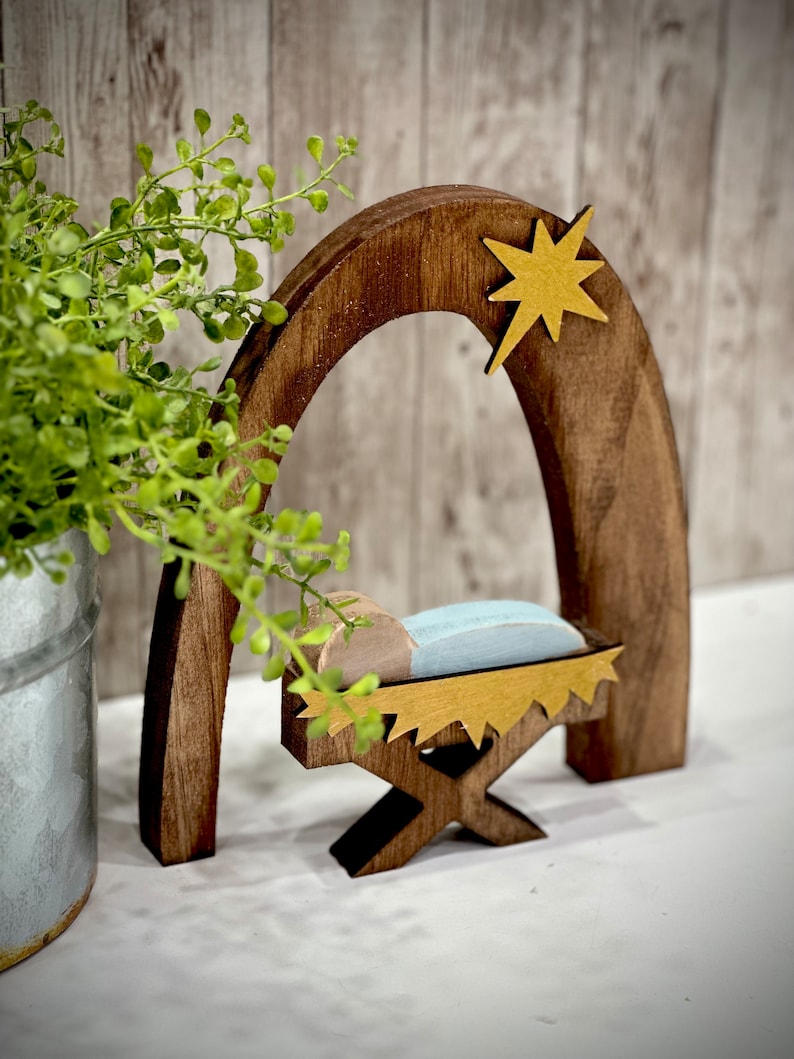 Op de afbeelding: Een houten kerststal met een donkerbruine boog en een gouden ster. Binnen de boog staat een wieg met een lichtblauw kussen en een houten baby. De wieg wordt ondersteund door een houten standaard. Een groene plant in een metalen pot staat op de achtergrond.