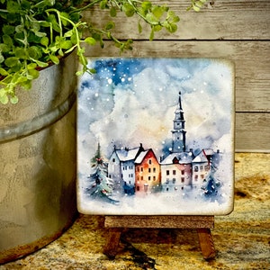 May include: A watercolor painting on a small wooden easel depicts a snowy village scene with a tall church steeple and several houses. The sky is a light blue with white snowflakes.