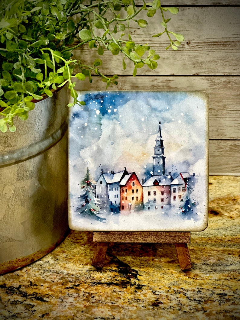 May include: A watercolor painting on a small wooden easel depicts a snowy village scene with a tall church steeple and several houses. The sky is a light blue with white snowflakes.