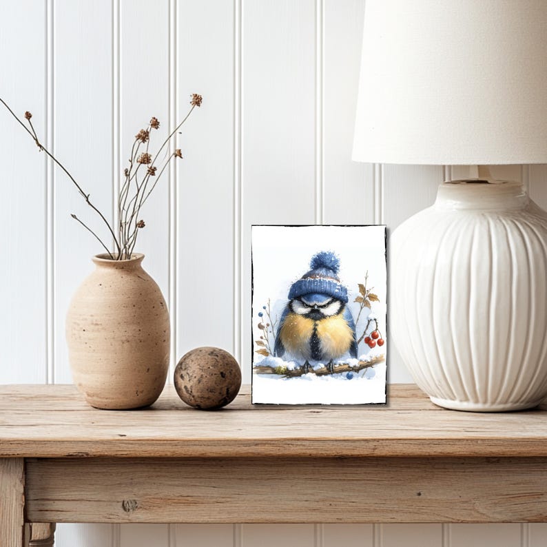 May include: A watercolor painting of a bluebird wearing a blue knit hat with a white pom pom. The bird is sitting on a branch with red berries and snow.