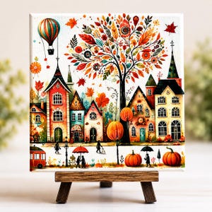 May include: A colorful autumn-themed artwork featuring a quaint village scene with houses, a large tree, and a hot air balloon. The artwork includes pumpkins, people with umbrellas, and a red trolley. The painting is displayed on a small wooden easel.