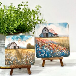 May include: Two small paintings on wooden easels, each depicting a white farmhouse with a red roof in a field of wildflowers. The painting on the left has a blue sky with white clouds, while the painting on the right has a blue sky with a few white clouds and a field of red, orange, and blue wildflowers.