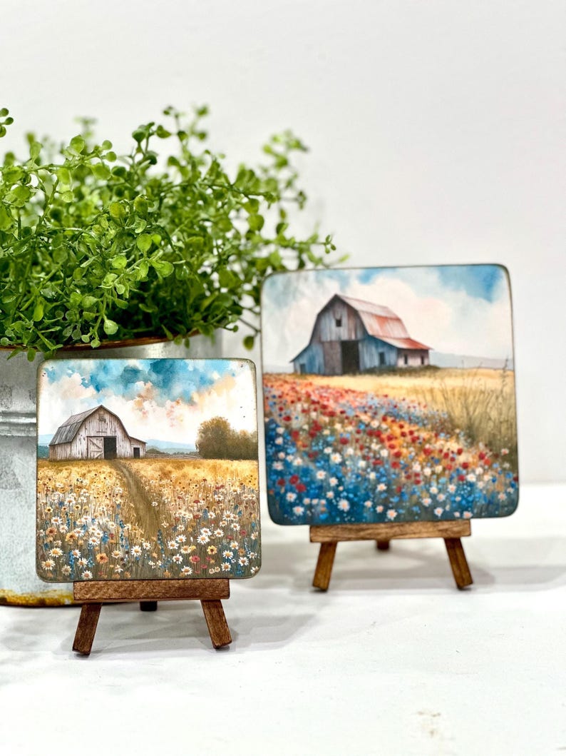 May include: Two small paintings on wooden easels, each depicting a white farmhouse with a red roof in a field of wildflowers. The painting on the left has a blue sky with white clouds, while the painting on the right has a blue sky with a few white clouds and a field of red, orange, and blue wildflowers.