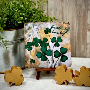 May include: A small square canvas painting with a gold background and green four-leaf clovers. The painting is on a small wooden easel and says "feeling lucky". There are three gold four-leaf clovers in the foreground.