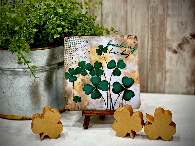 May include: A small square canvas painting with a gold background and green four-leaf clovers. The painting is on a small wooden easel and says "feeling lucky". There are three gold four-leaf clovers in the foreground.