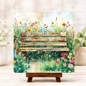 May include: A square decorative print featuring a watercolor painting of a wooden park bench surrounded by colorful wildflowers. The bench has the names "Lili + Jayden" inscribed on it. The print is displayed on a small wooden easel.