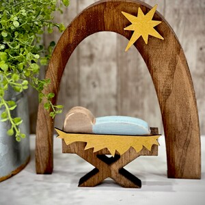 Op de afbeelding: Houten kerststal met een baby in een kribbe, onder een boog en een ster. De baby heeft een lichtblauwe deken en een bruin hoofd. De boog en de ster zijn goudkleurig. De kribbe is goud en bruin.
