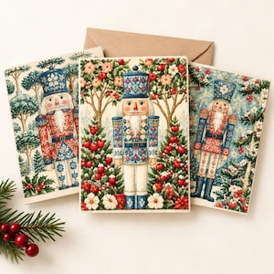 May include: Three greeting cards featuring a nutcracker illustration in blue, red, and white. Each card displays a nutcracker figure standing among trees with red berries and white flowers. A brown envelope is positioned behind the cards.