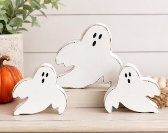 May include: Three white wooden ghost figures with distressed edges and black painted eyes and mouths. The ghosts are shaped with rounded heads and flowing, arm-like extensions, arranged in a group with one larger ghost centered behind two smaller ones.