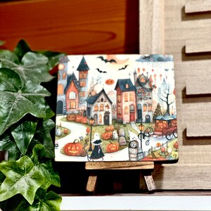 May include: A square Halloween-themed artwork on a small wooden easel. The artwork depicts a whimsical town scene with houses, pumpkins, ghosts, and bats under a full moon. The color palette includes orange, black, and various other colors.
