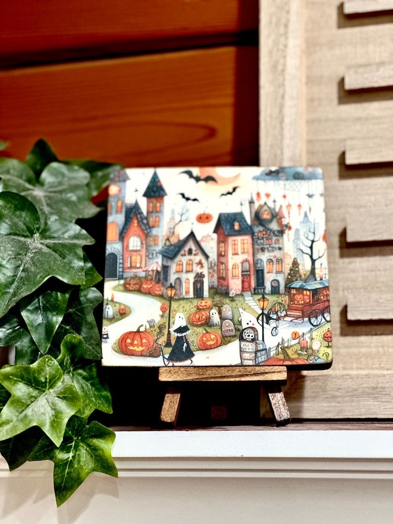 May include: A square Halloween-themed artwork on a small wooden easel. The artwork depicts a whimsical town scene with houses, pumpkins, ghosts, and bats under a full moon. The color palette includes orange, black, and various other colors.