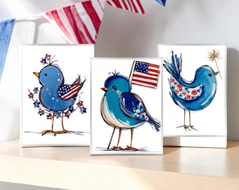 May include: Three square art prints featuring whimsical blue birds with patriotic themes. One bird has an American flag wing, another holds a flag, and the third has floral accents. Red, white, and blue bunting hangs in the background.