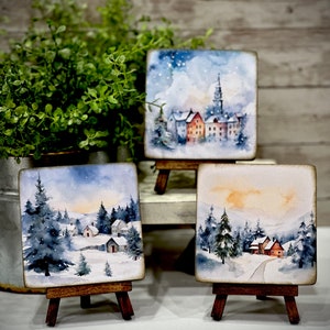 May include: Three small watercolor paintings on easels depicting winter scenes. The paintings feature snow-covered houses, trees, and a snowy landscape. The paintings are framed in wood and have a rustic look.