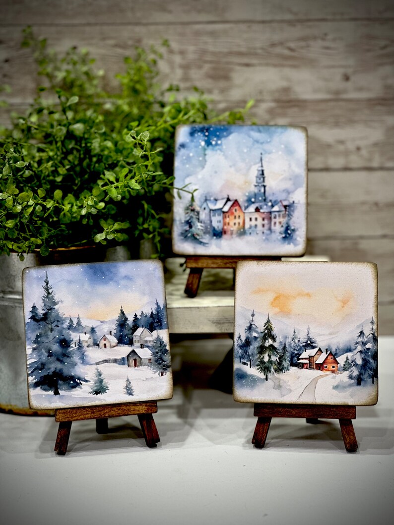 May include: Three small watercolor paintings on easels depicting winter scenes. The paintings feature snow-covered houses, trees, and a snowy landscape. The paintings are framed in wood and have a rustic look.