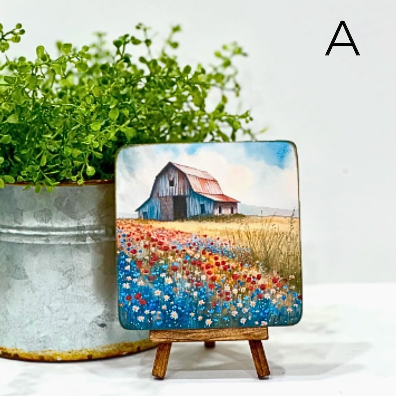 May include: A small painting on a wooden easel depicts a red barn with a blue roof in a field of wildflowers. The flowers are blue, red, and white. The barn is in the distance, and the field of flowers is in the foreground.