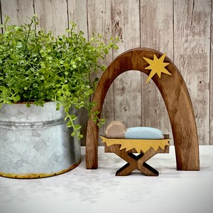 Op de afbeelding: Houten kerststal met een ster, boog en kribbe. De kribbe bevat een babyfiguur met een blauwe deken. Een groene plant in een metalen emmer staat links. De achtergrond is een licht houten paneelwand.