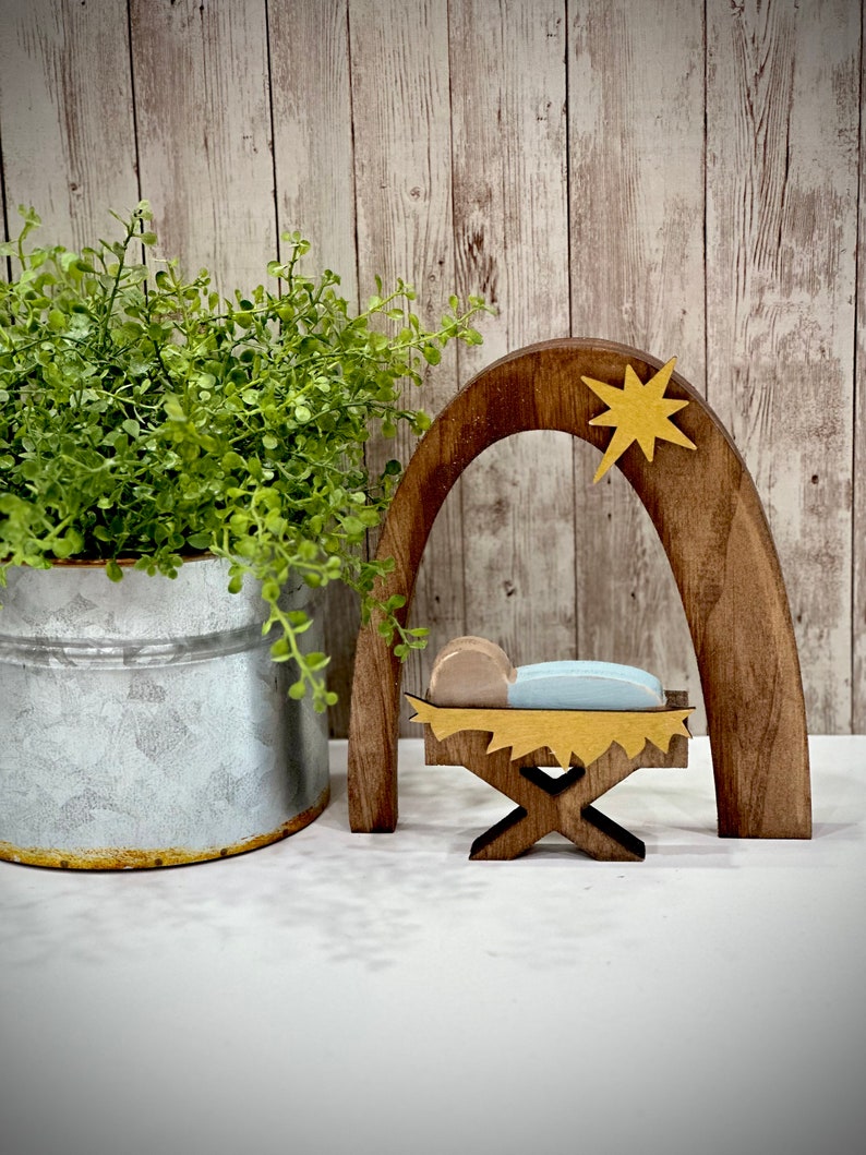 Op de afbeelding: Houten kerststal met een ster, boog en kribbe. De kribbe bevat een babyfiguur met een blauwe deken. Een groene plant in een metalen emmer staat links. De achtergrond is een licht houten paneelwand.