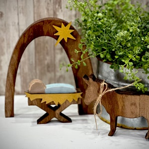 Op de afbeelding: Houten kerststal met een ster, boog en kribbe. Een houten ezel met een touwstrik staat in de buurt. Een metalen emmer met groen gebladerte staat op de achtergrond. De sc&egrave;ne staat op een wit oppervlak.